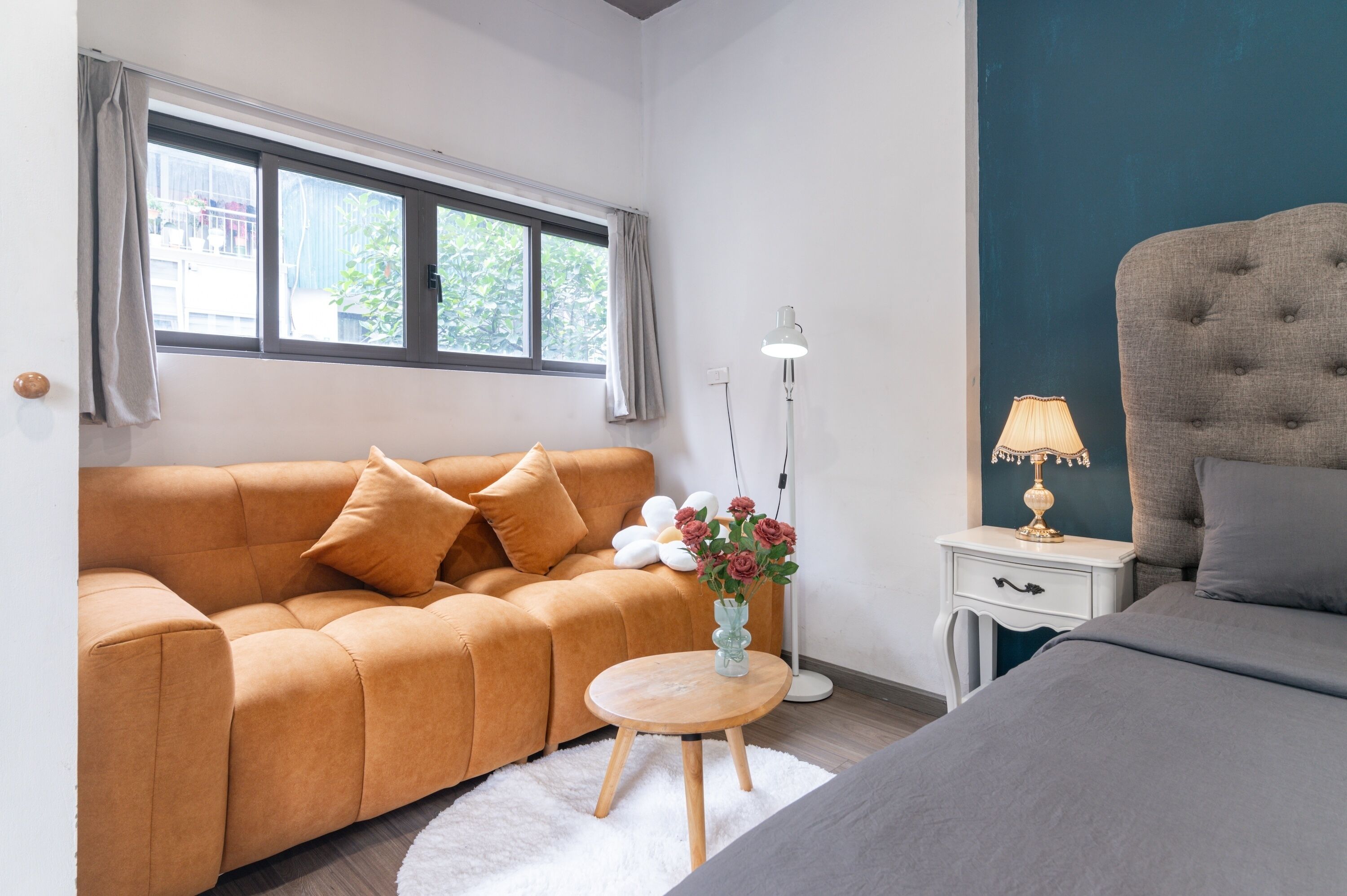 A modern room with a sofa, a bedside table with a lamp, a small round table with a vase of flowers, and a bed.
