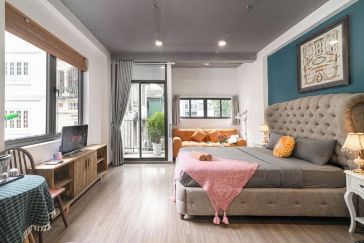 A modern bedroom with a large bed, a TV on a wooden stand, a dining table, and a view through a glass door.