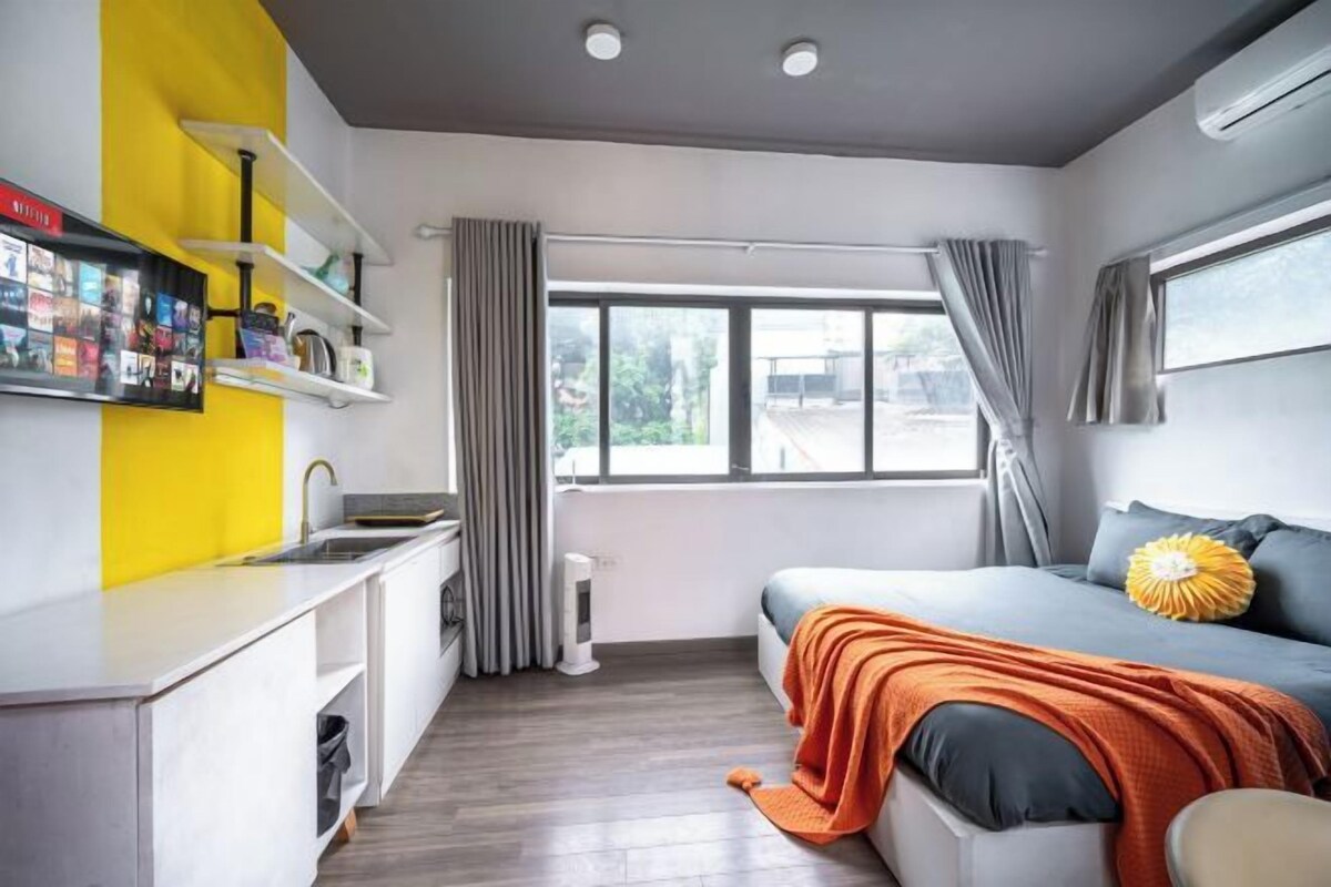 A compact room with a kitchenette, a bed with an orange blanket, a window with curtains, and a shelf with items.