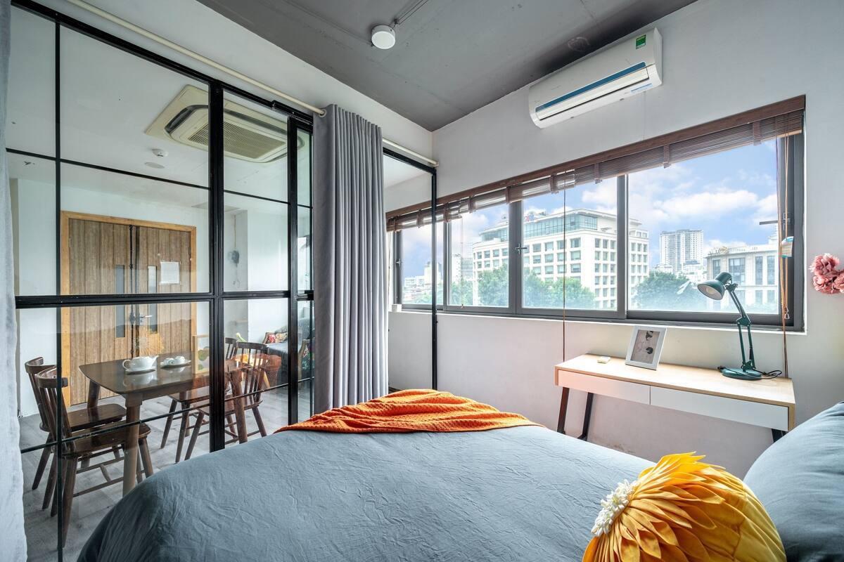 A modern hotel room with a large window, a desk with a lamp, a bed with a blanket, and a dining area with chairs and a table.