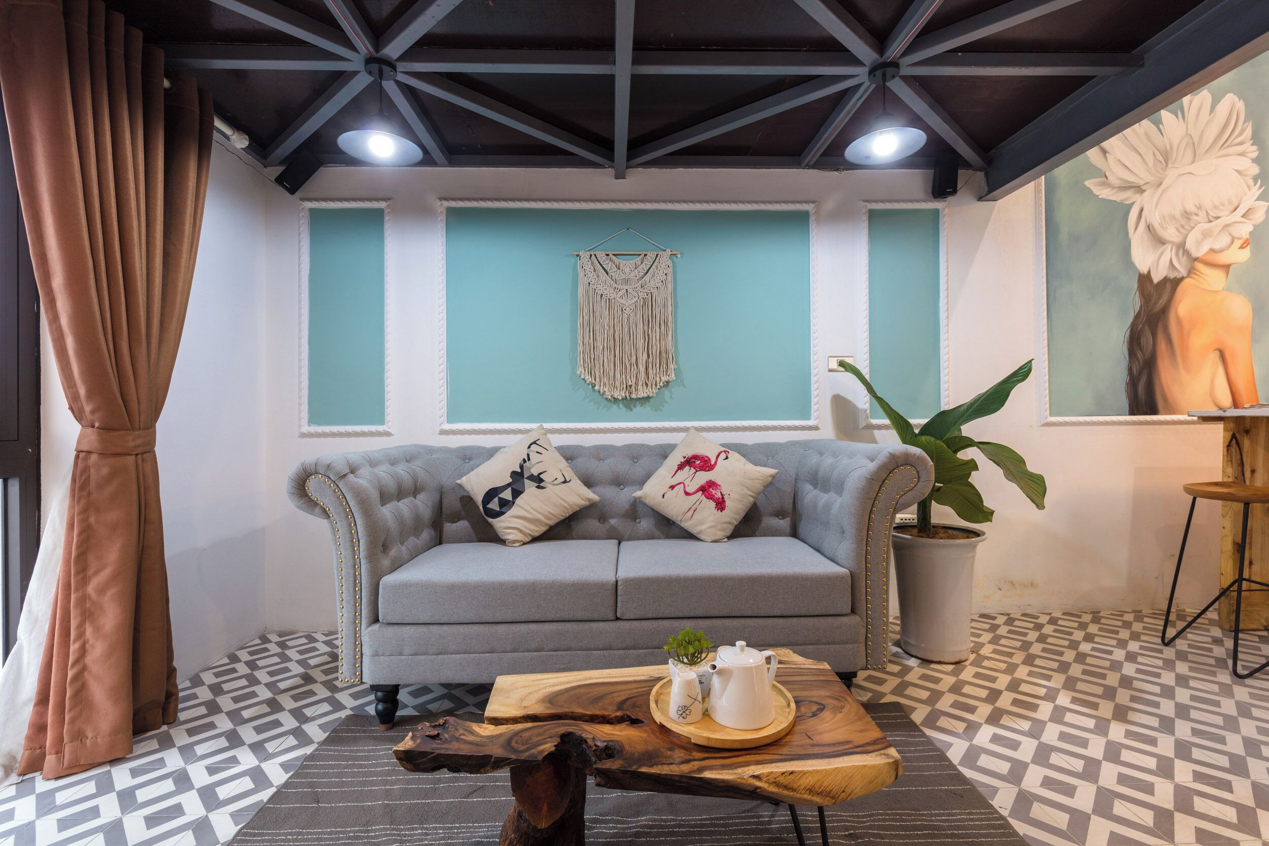 Ein Wohnzimmer mit grauem Sofa, einer Wanddekoration aus Makramee, einer Topfpflanze und einem Holztisch mit Teekanne und Tasse.