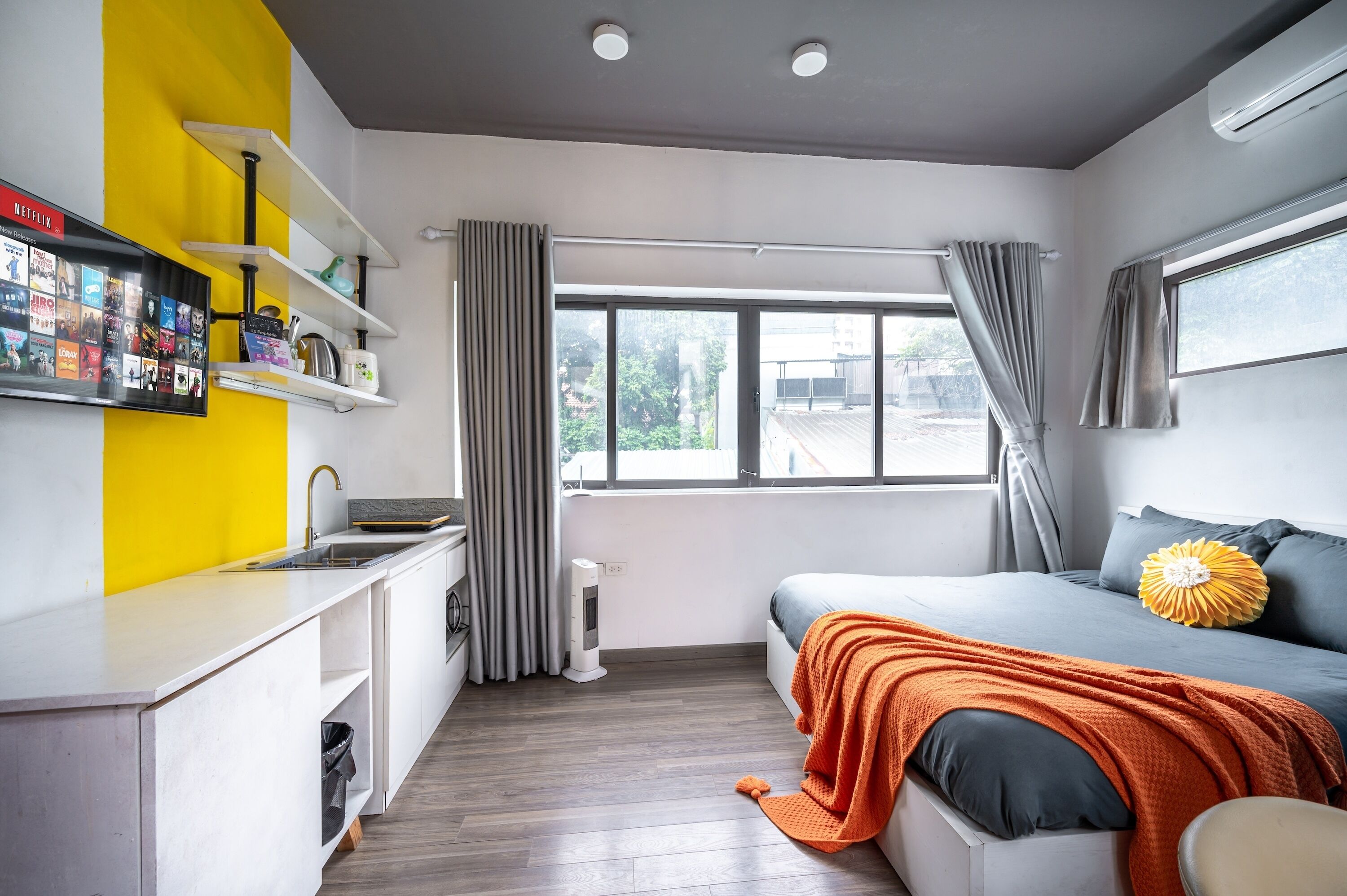 A compact hotel room with a kitchenette, a bed with an orange blanket, and a large window with curtains.