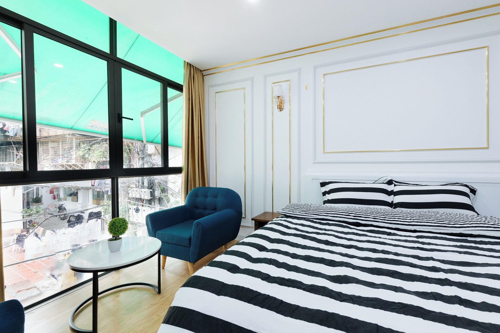 A hotel room with a large window, a bed with striped bedding, a blue armchair, and a small table with a plant.