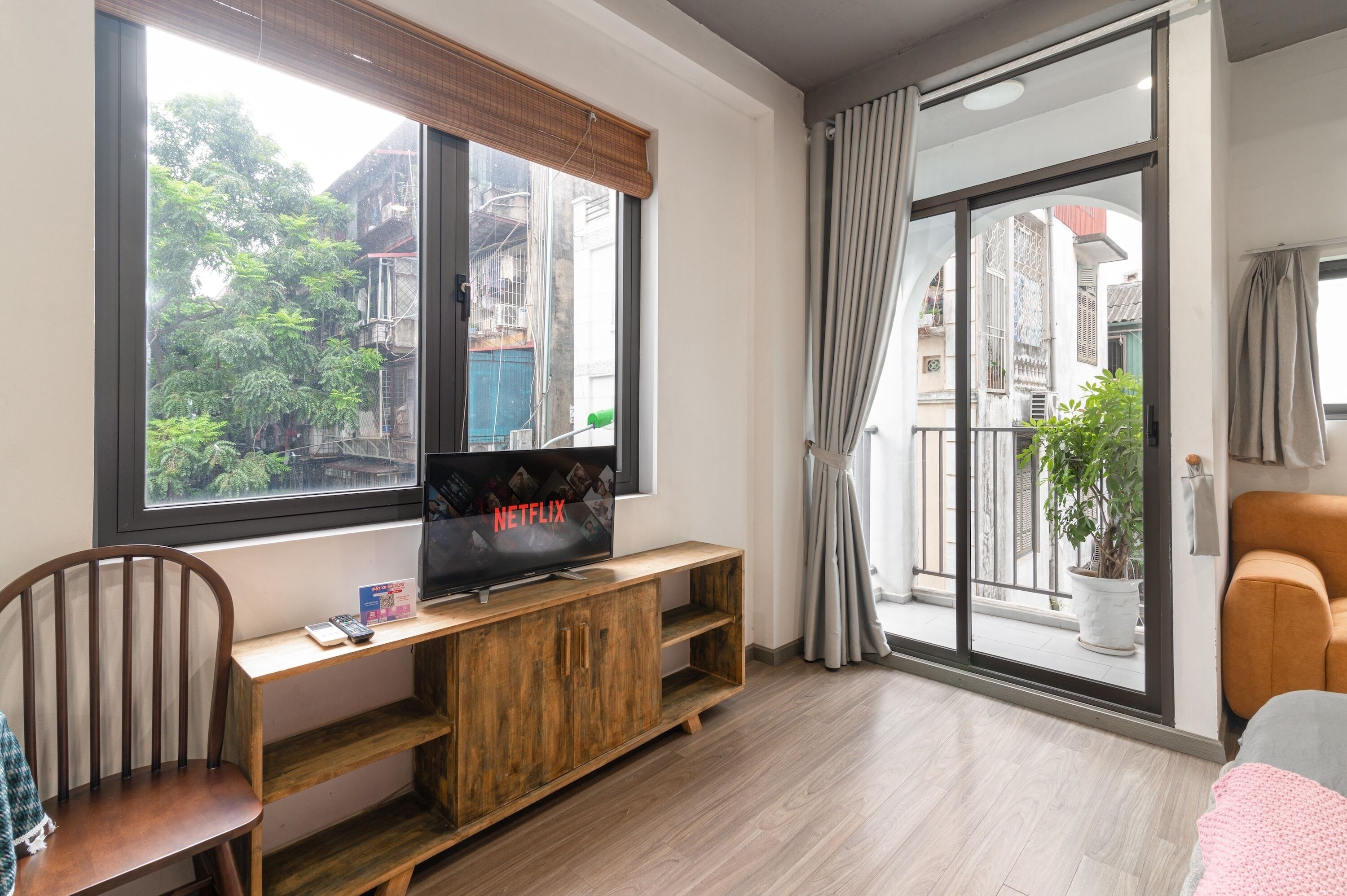 Een woonkamer met een houten tv-meubel, een tv waarop 'NETFLIX' staat, een fauteuil, een bank, een raam met uitzicht op gebouwen en bomen, en een schuifdeur met glas.