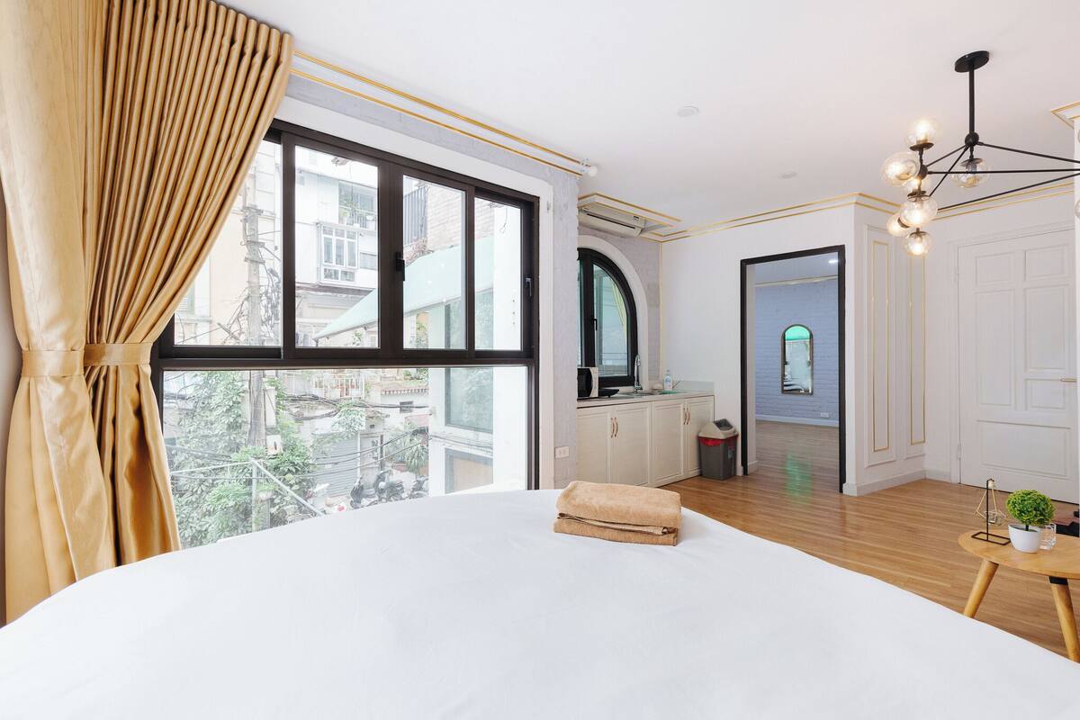 A hotel room with a large bed, a view of the city, a bathroom with a mirror, and a small table with a plant.