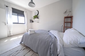A bedroom with a bed, a bedside table, a wall clock, a shelf with a plant, and a window with curtains. - Artistic Vac Villa w pool by Sea N' Rent (Rishon LeTsiyon)