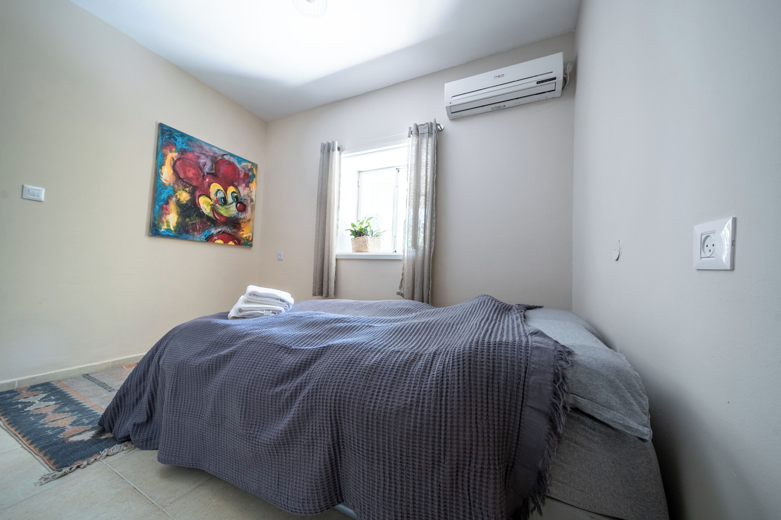 A bedroom with a bed, a window with curtains, an air conditioner, and a painting on the wall.