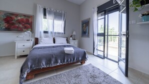 A bedroom with a bed, a nightstand, a lamp, a painting, and a view through a glass door. - Artistic Vac Villa w pool by Sea N' Rent (Rishon LeTsiyon)