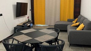 Ein modernes Wohnzimmer mit einem grauen Sofa, gelben Vorhängen, einem Flachbildfernseher und einem Essbereich mit Tisch und Stühlen.