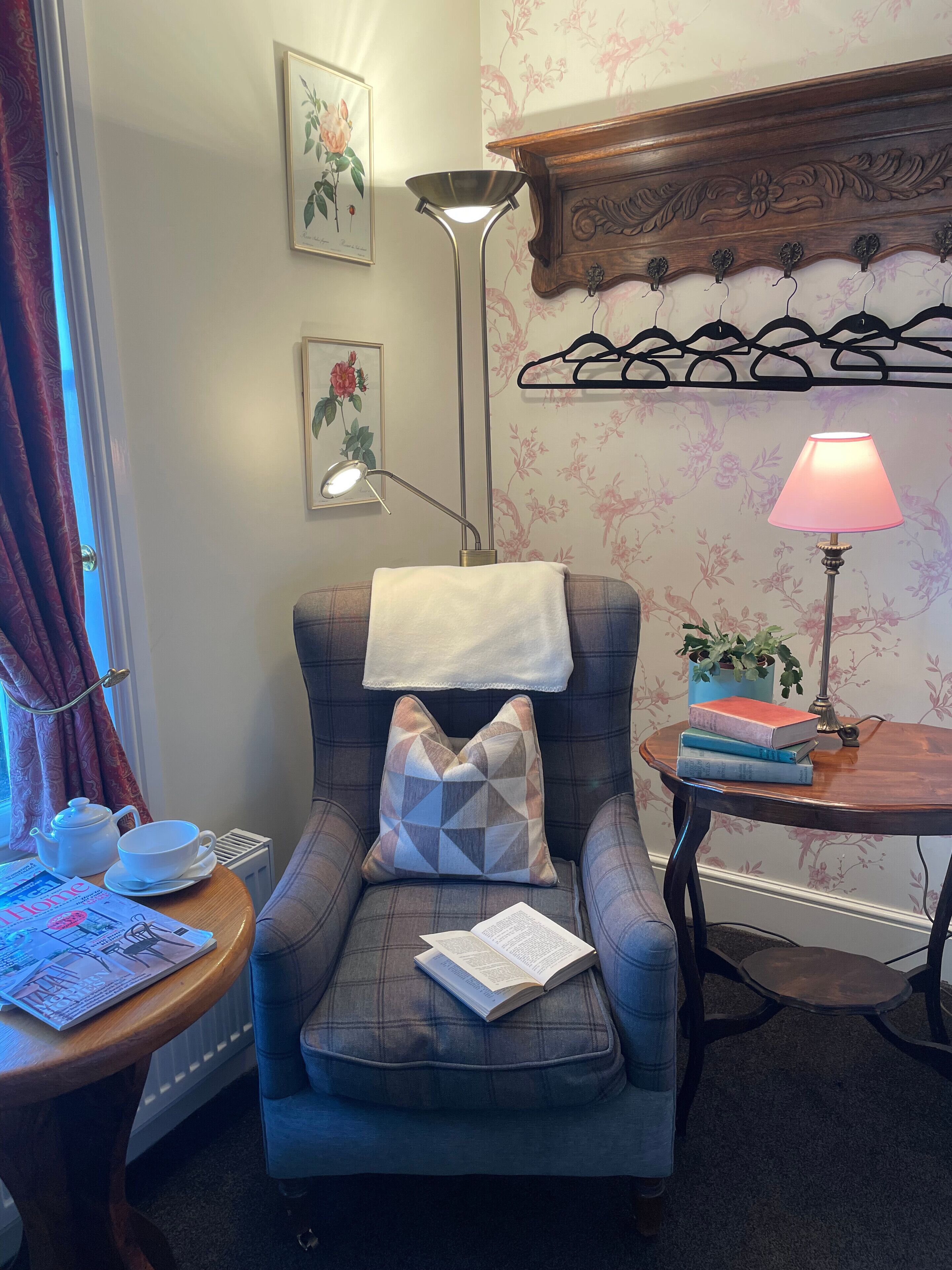 Een knus ingerichte kamer met een geruit fauteuil, een klein houten tafeltje met een lamp, een radiator en een raam met gordijnen.