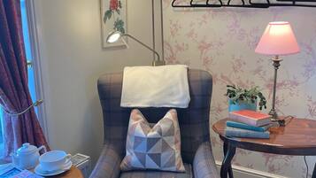 Een knus ingerichte kamer met een geruit fauteuil, een klein houten tafeltje met een lamp, een radiator en een raam met gordijnen.