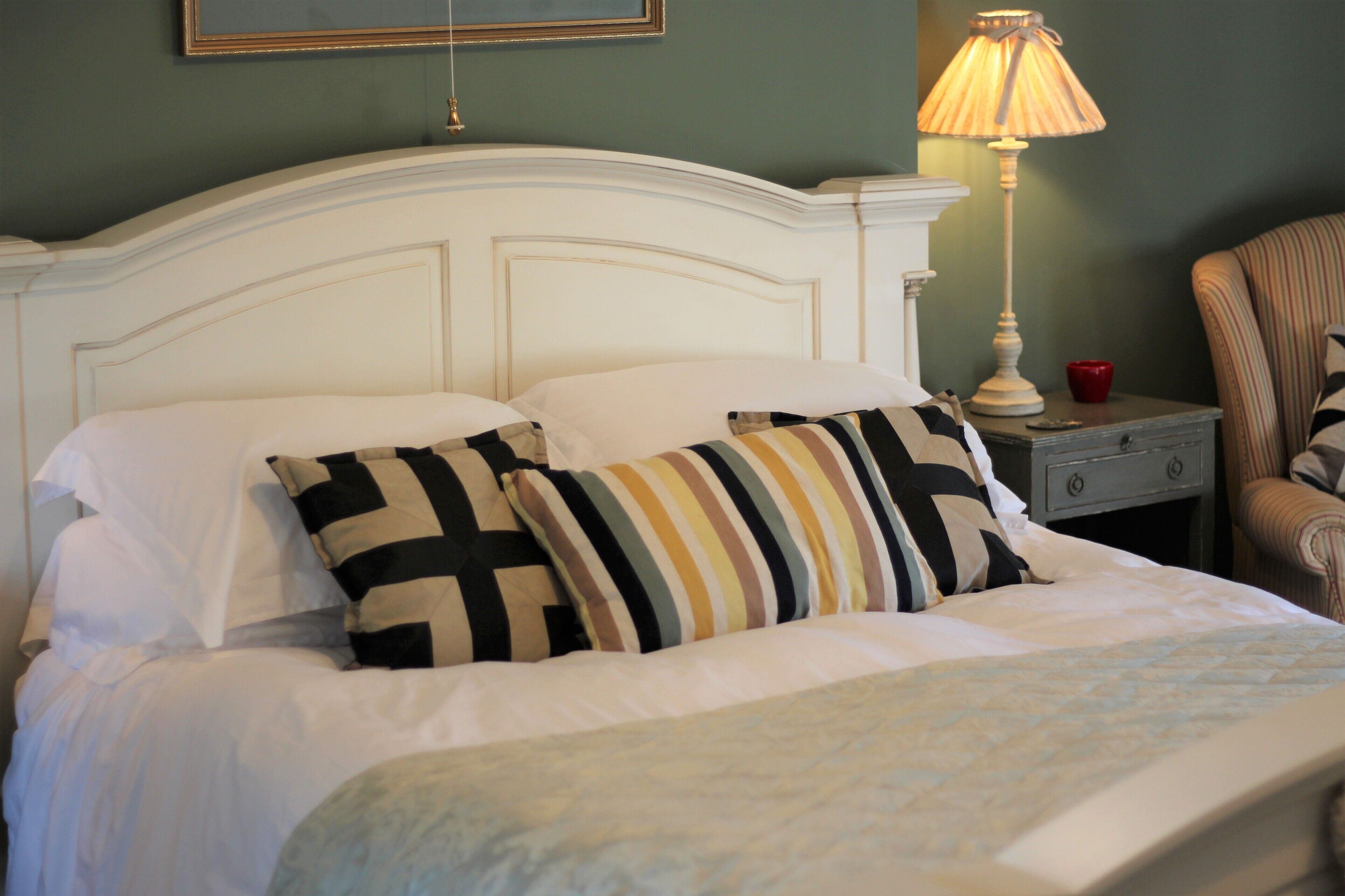 A neatly made bed with striped pillows, a bedside lamp, and a chair in a room with green walls.