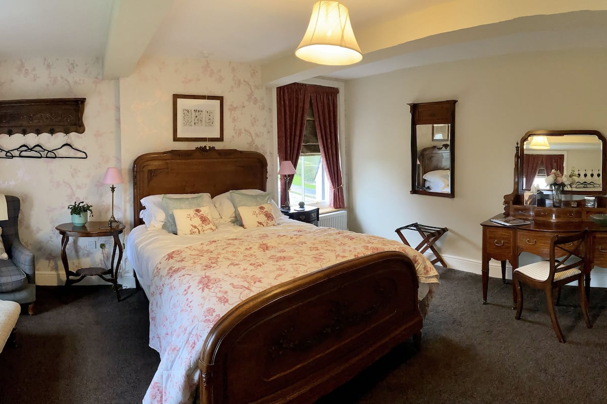 A bedroom with a large bed, a wooden dresser, a chair, a small table with a lamp, and a window with curtains.