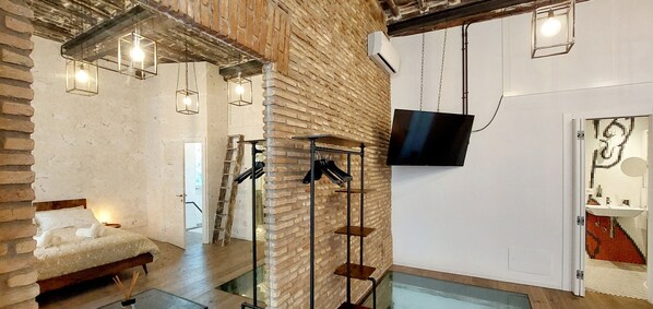 A modern room with a brick wall, a bed with a white duvet, a TV mounted on the wall, and a small table with a lamp.