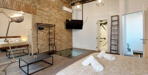 Habitación de hotel moderna con pared de ladrillo, televisor de pantalla plana, ducha de vidrio, piso de madera, cama con colcha blanca, escritorio con lámpara y escalera cerca de la puerta.