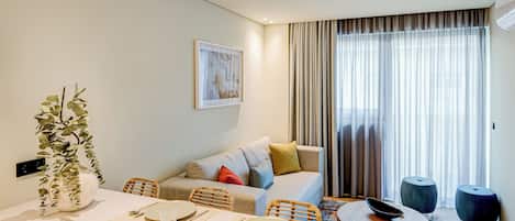 A modern hotel room with a dining area, a sofa, and a large window with curtains.