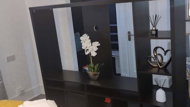 Una camera da letto con un letto, una coperta gialla, una cassettiera con uno specchio, una pianta in vaso e una decorazione a forma di cuore.