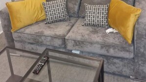 Un soggiorno con un divano grigio, due cuscini gialli, un tavolino in vetro e un frigorifero nero.