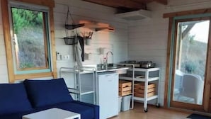 L’intérieur d’un chalet accueillant, avec un canapé bleu, une petite table, une chaise en bois, un coin cuisine et de grandes fenêtres.