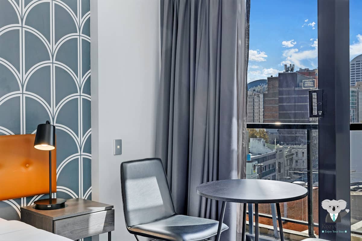 A modern hotel room with a city view, a round table, a chair, and a lamp.