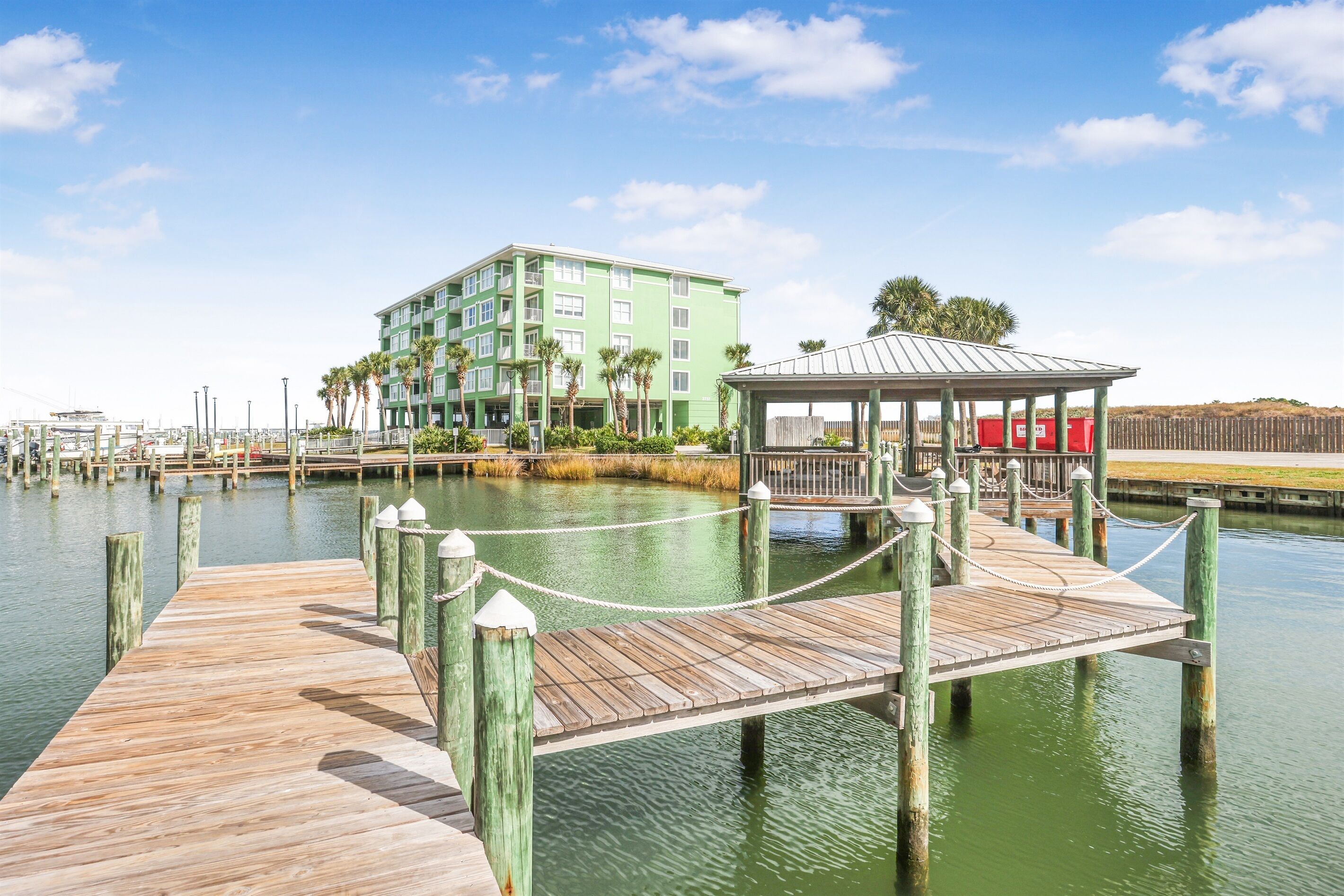 A wooden dock leading to a gazebo over water, with a green building in the background.
