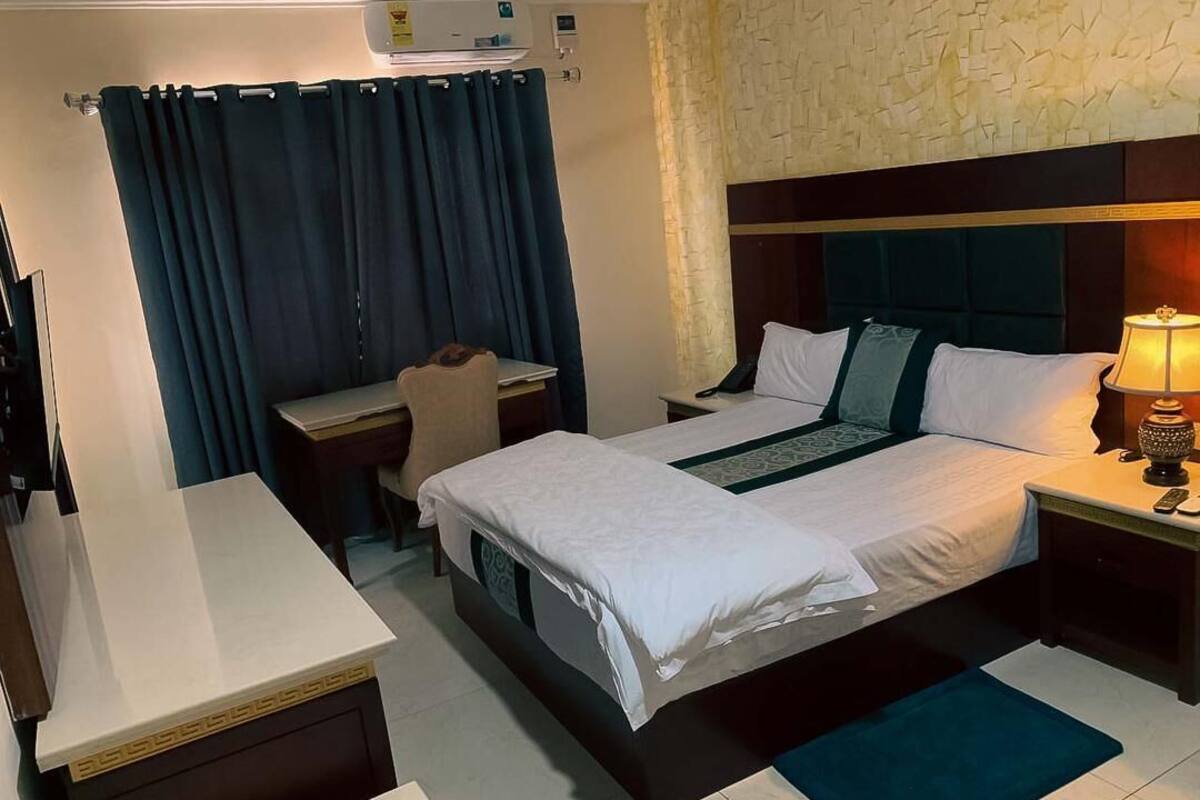 A hotel room with a bed, desk, chair, and a nightstand with a lamp.