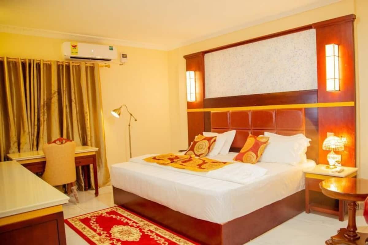 A hotel room with a large bed, a desk, a chair, a red armchair, and a small table with a lamp.