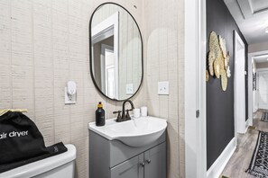 A bathroom with a white sink, a round mirror, and a black hair dryer bag on the toilet. - Boho 3BR Near Mercedes-Benz Stadium & BeltLine | Pet-Friendly (Atlanta)