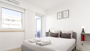 Um quarto moderno com uma cama grande, uma mesa de cabeceira com uma luminária, uma pequena estante com livros e uma janela com vista para prédios.