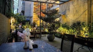 Un espacio exterior cubierto para comer con mesas y sillas, rodeado de plantas en macetas y un árbol grande.