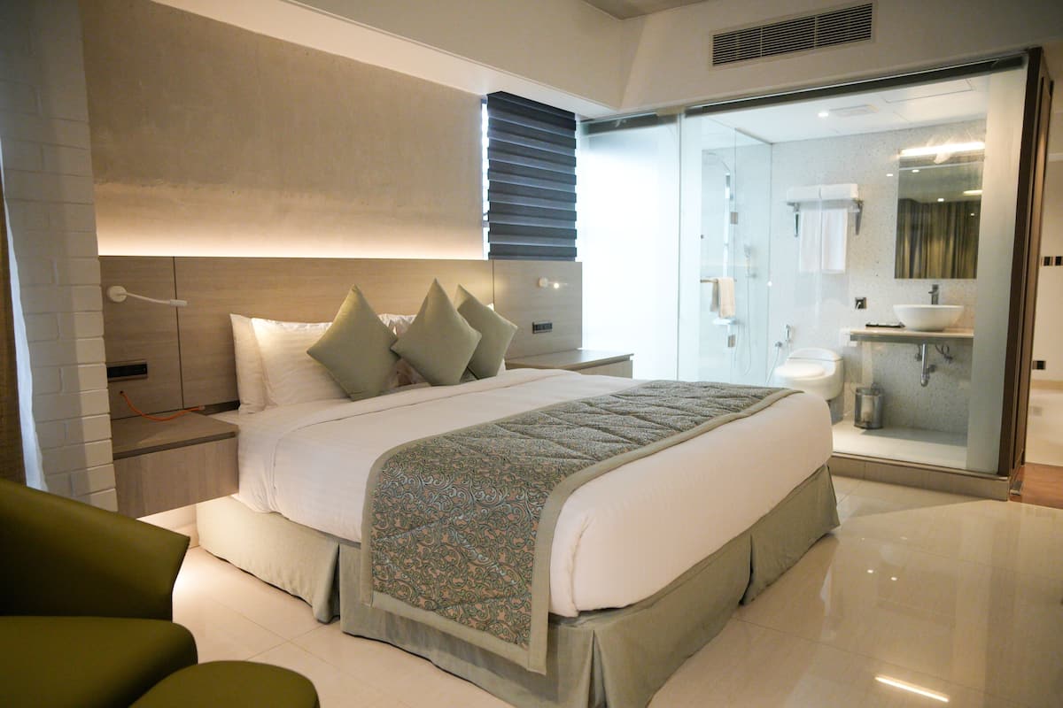 A modern bedroom with a large bed, a nightstand, a green armchair, and a glass-enclosed bathroom with a sink and mirror.