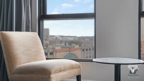 A room with a view of a cityscape through a large window, a chair, and a small round table. - KozyGuru | Sydney CBD | 2 Bed Studio (Haymarket)