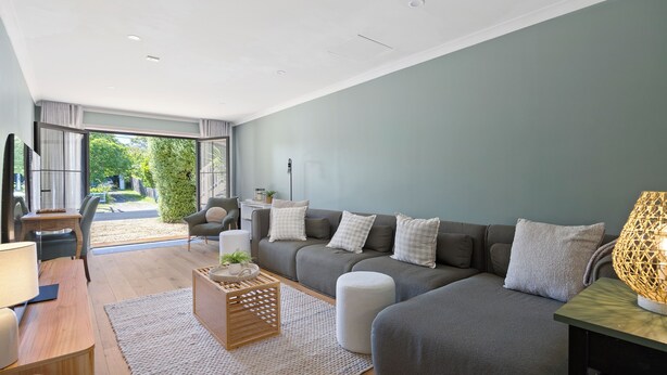 A modern living room with a grey sofa, a wooden coffee table, and a large window offering a view of greenery outside. - Rosebud Township Entertainer (Melbourne)