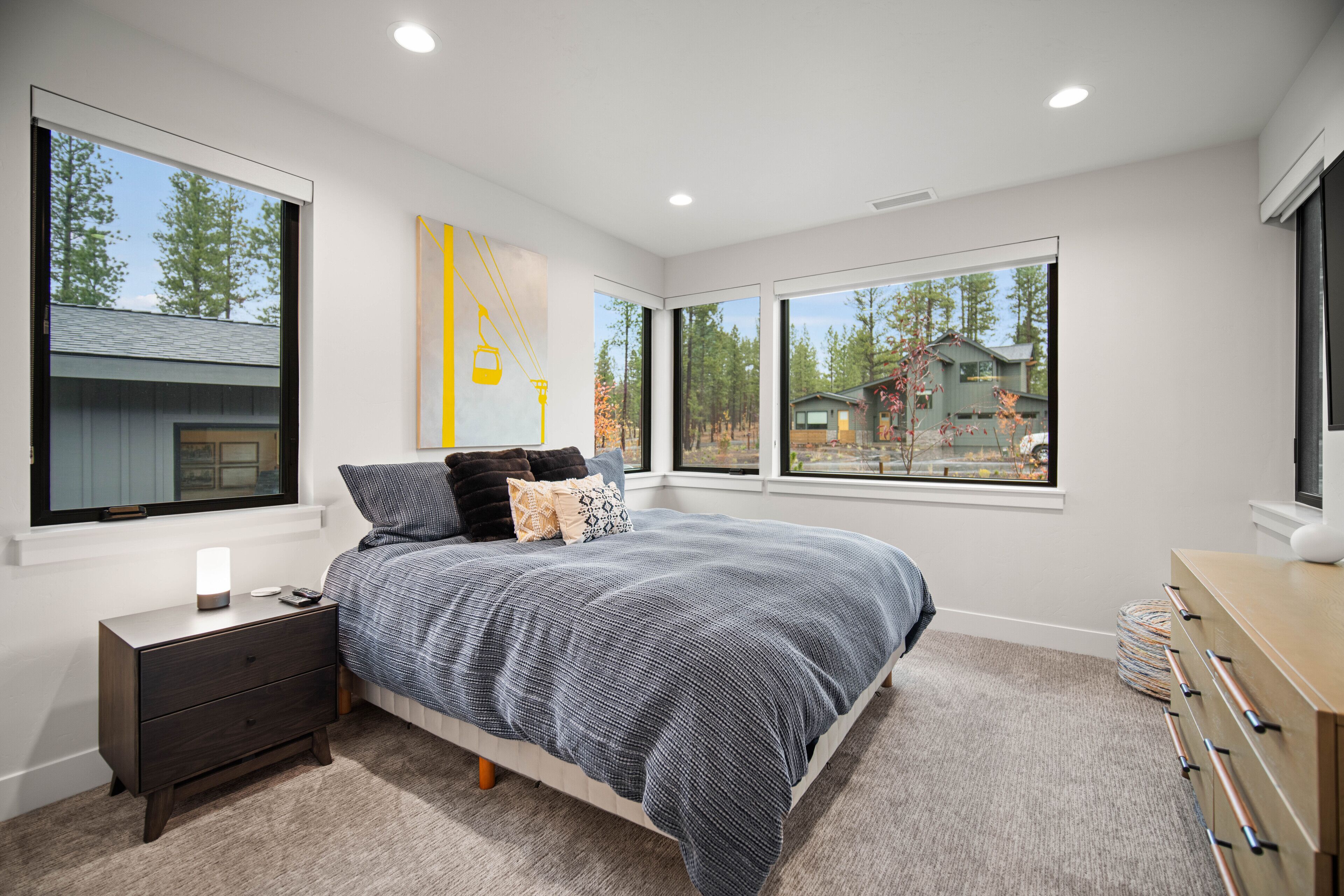 A bedroom with a large bed, a nightstand, a dresser, and multiple windows offering views of the outdoors.