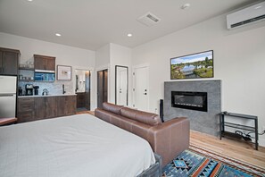 A modern bedroom with a large bed, a seating area, a fireplace, and a kitchen in the background. - Designer Studio with Access to Forest House (Bend)