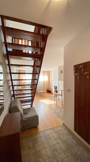 Una escalera con barandales de madera que sube a un nivel superior, un rincón de descanso con un banco y una mesa, y una puerta con ganchos.