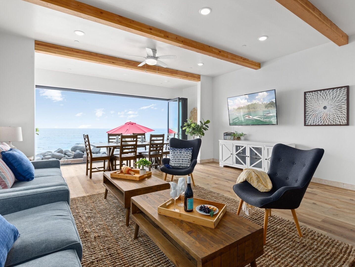 A spacious living room with a large window overlooking the ocean, wooden furniture, and a modern ceiling fan.