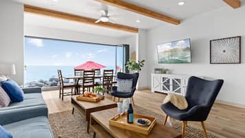 A spacious living room with a large window overlooking the ocean, wooden furniture, and a modern ceiling fan.