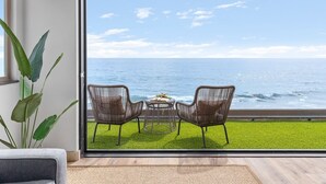 A room with a large sliding glass door leading to a balcony with a view of the ocean. There are two wicker chairs and a small table on the balcony.