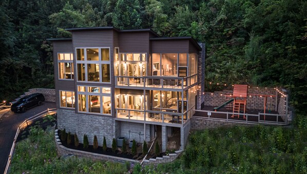 A multi-level modern house with large windows, a stone foundation, and a landscaped garden with a play area. - EXTRAORDINARY Contemporary Luxury Cabin! Screened Porches, Fireplaces, WOW View! (Gatlinburg)