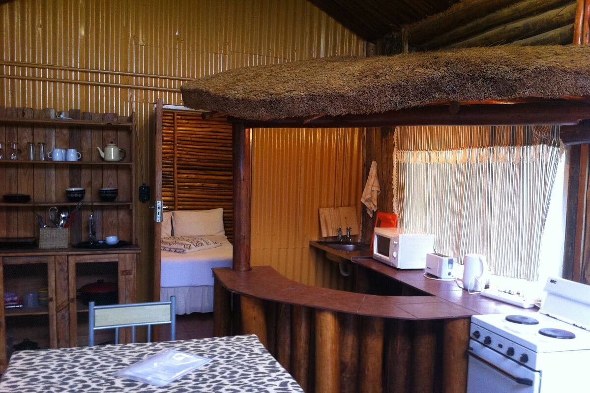 A rustic kitchen with wooden walls, a bed with a patterned bedspread, a kitchenette with a microwave and stove, and a dining area with a table and chairs.