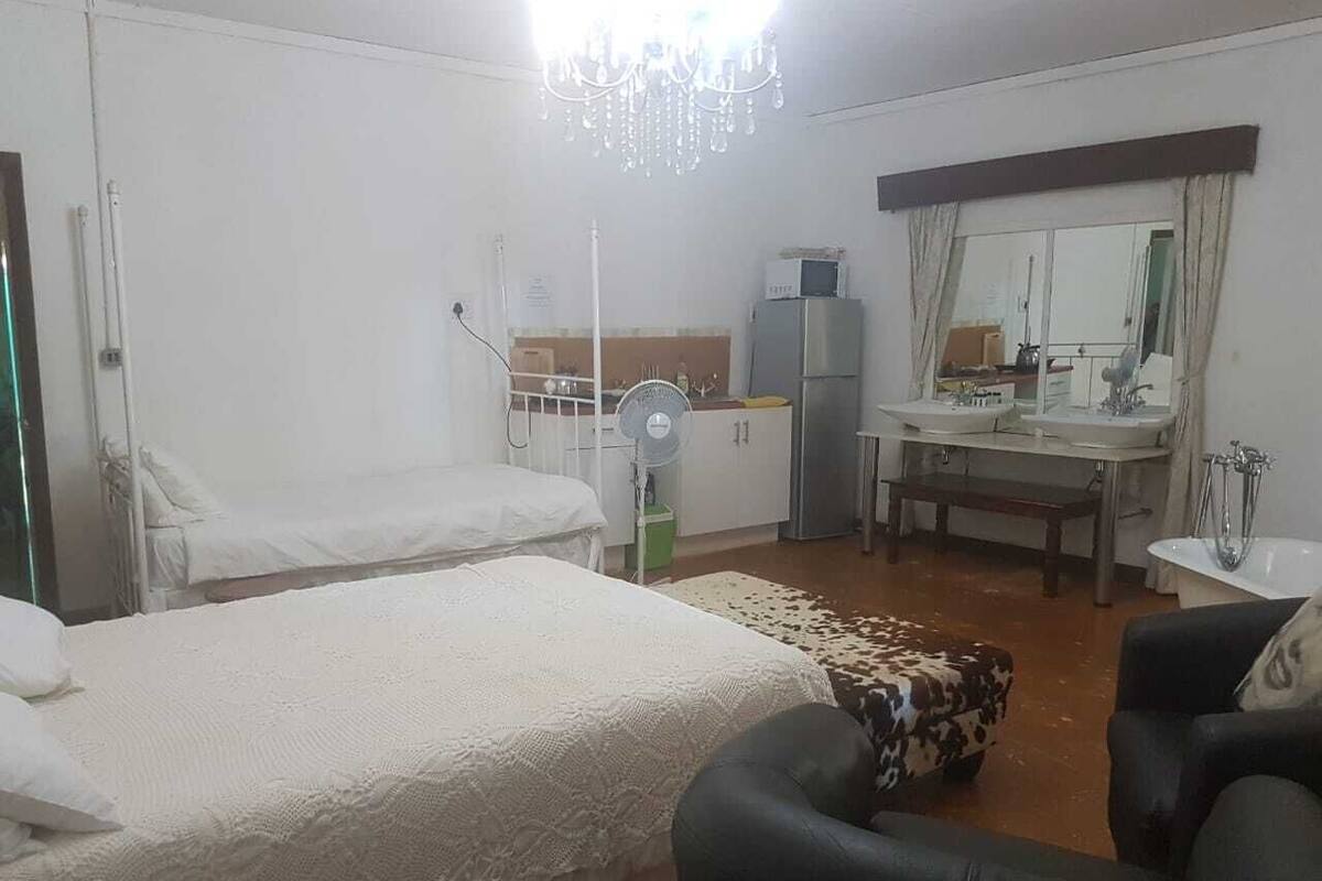 A hotel room with two beds, a sitting area with a chair, a small table, a mirror, a window with curtains, and a kitchenette with a refrigerator and microwave.