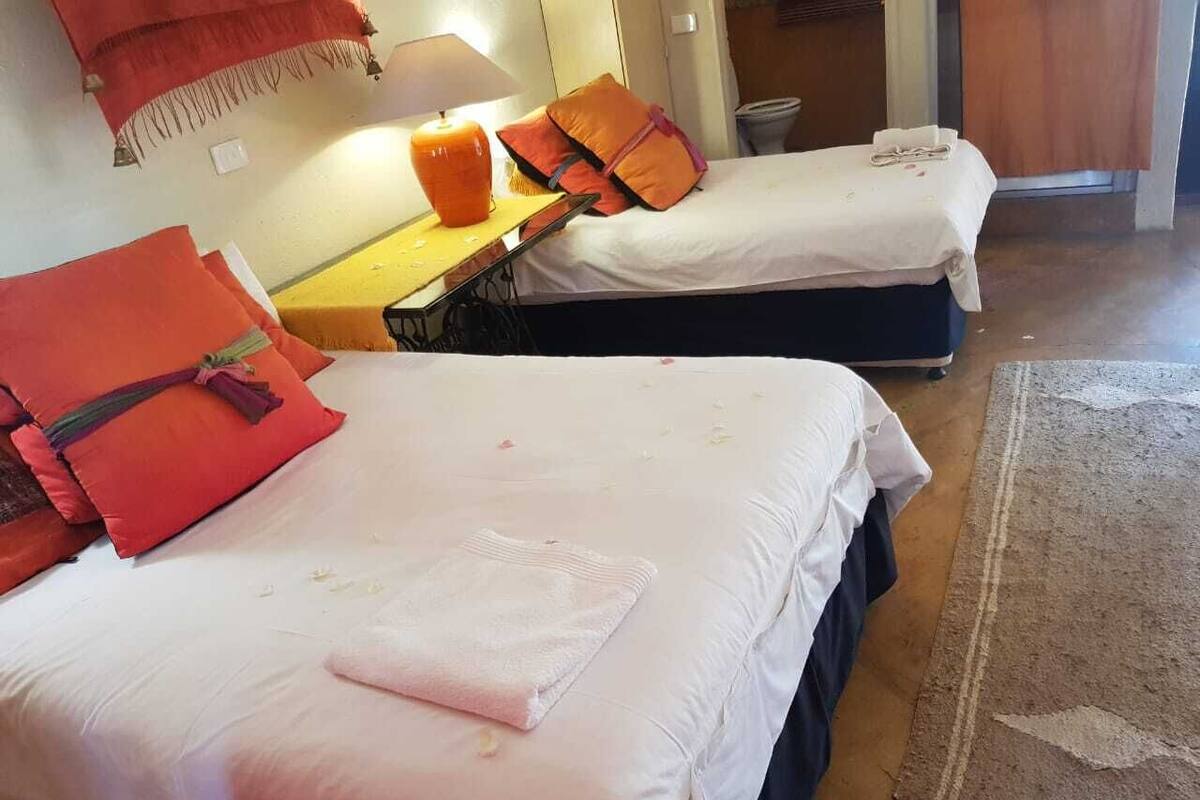 A hotel room with two beds, each with a white mattress and orange pillows. There is a small table with a lamp and a few items on it.
