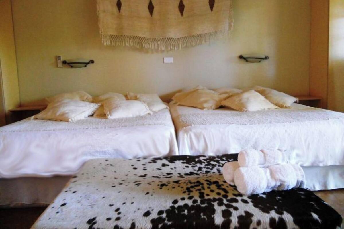 A room with two beds, each with multiple pillows and a blanket. There is a small table and a chair in the corner.