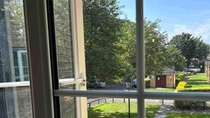 Blick durch ein Fenster mit Metallrahmen, ein gepflasterter Weg und ein kleines Gebäude mit einer Tür.