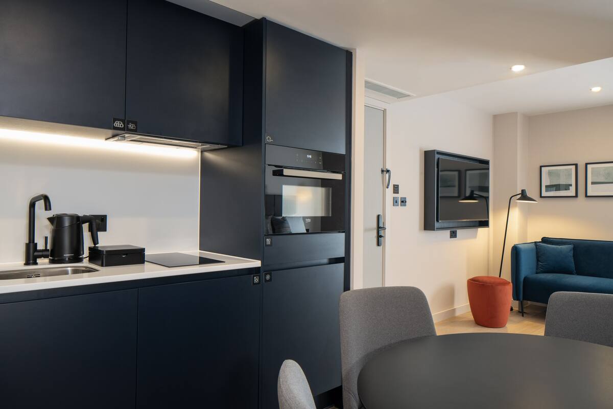 Eine moderne Küche mit dunklen Schränken, einem eingebauten Ofen und einer Kaffeemaschine. Ein Essbereich mit Tisch und Stühlen lädt zum Verweilen ein, im Hintergrund befindet sich ein blaues Sofa.