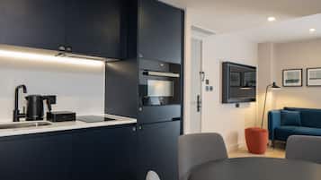 A modern kitchen with dark cabinetry, a built-in oven, and a coffee machine. There is a dining area with a table and chairs, and a blue sofa in the background.