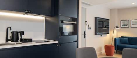 Una cocina moderna con armarios oscuros, horno empotrado y cafetera. Hay un comedor con mesa y sillas, y un sofá azul al fondo.
