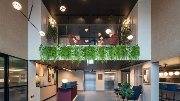 Un espacio de oficina moderno con una recepción, dos ascensores, un área de descanso con sillas y un balcón con plantas.