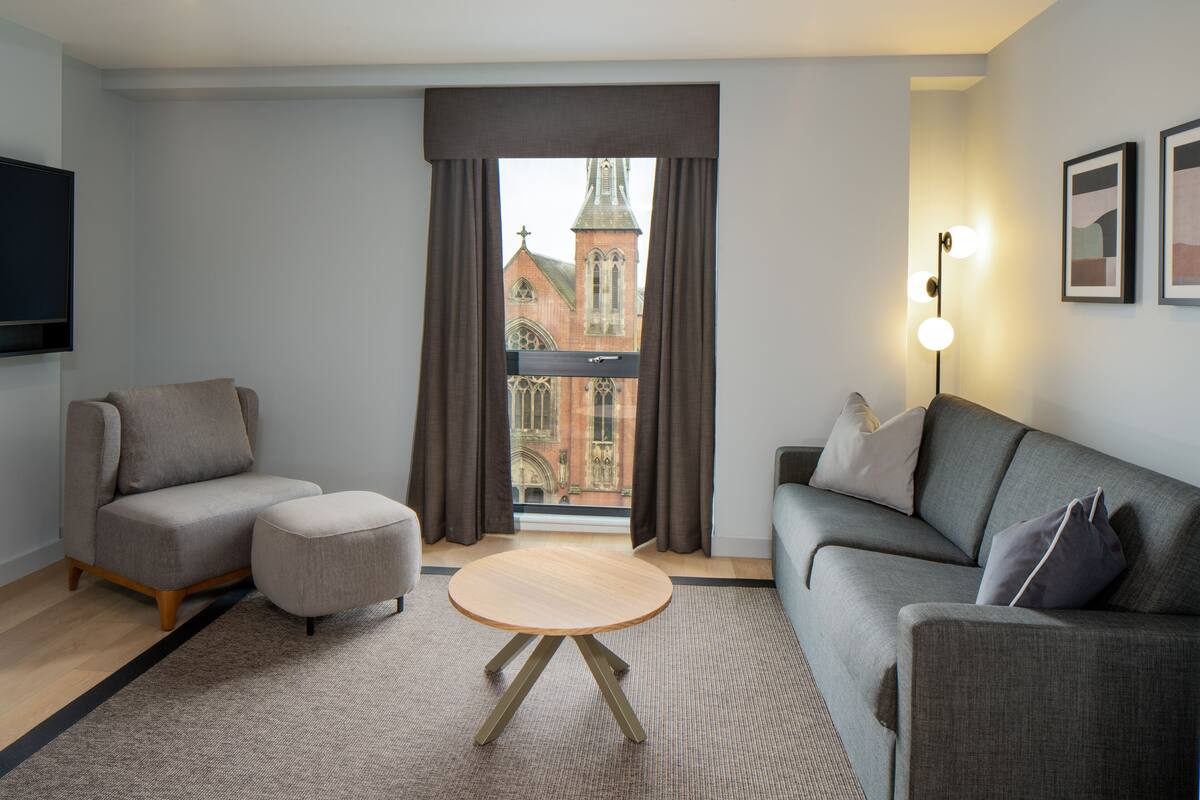 Ein modernes Wohnzimmer mit einem grauen Sofa, einem runden Holztisch und einem grauen Sessel. Ein großes Fenster mit Vorhängen bietet Blick auf ein Gebäude mit einem Turm.
